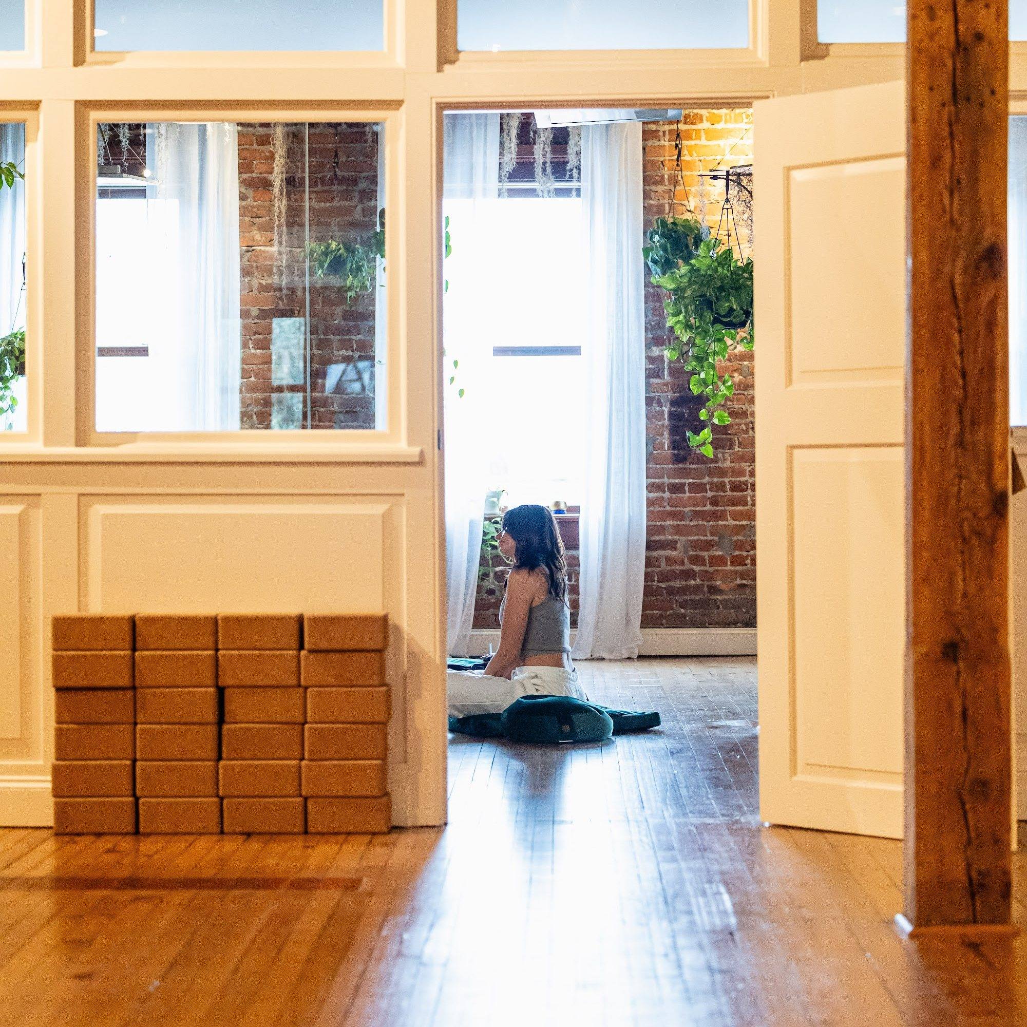 A stack of cork blocks by the doorway while a student meditates in the sunlit studio
