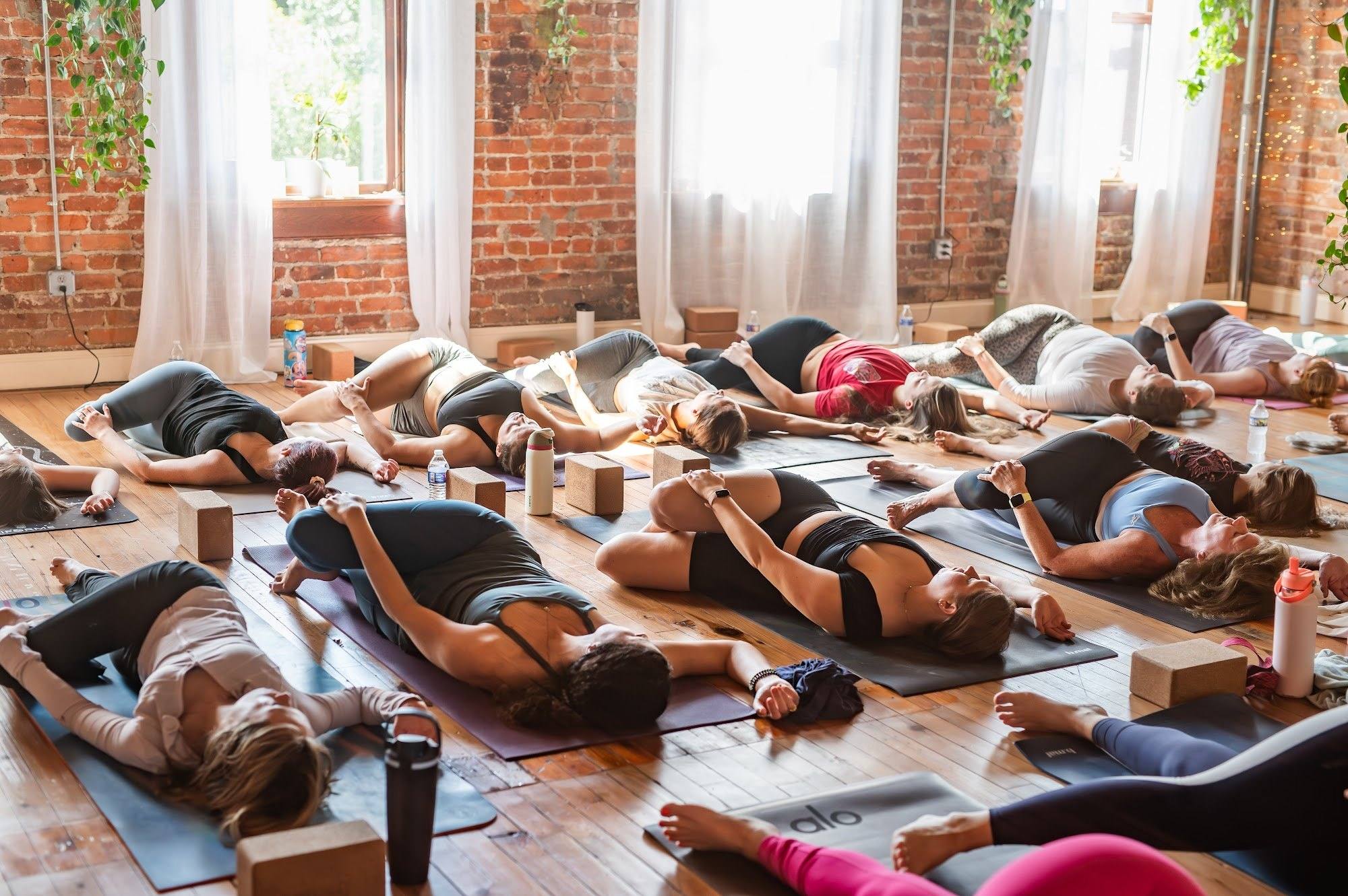 A full class resting in savasana on their mats in the sunlit brick studio at The Yoga Loft